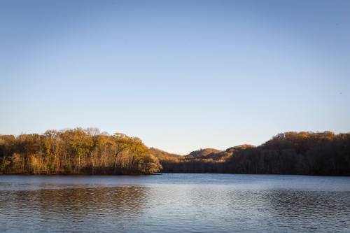 A serene lake surrounded by trees and hills under a clear blue sky.