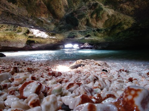 A rocky cave by the sea, filled with smooth, colorful shells and sunlight reflecting on the water.