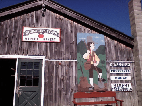 A rustic wooden building with signs for a market and bakery, featuring a painted figure carrying a backpack.