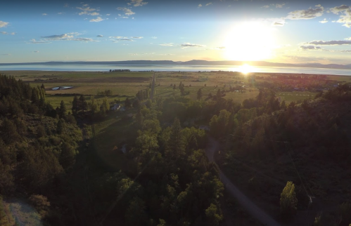 A scenic landscape at sunset, featuring a lake, fields, and trees under a colorful sky.