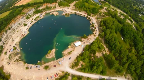 Aerial view of a circular quarry lake surrounded by trees and a sandy shore, with boats and vehicles nearby.