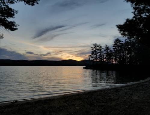 A serene lakeside view at sunset, with silhouettes of trees and a colorful sky reflecting on the water.