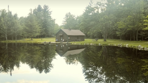 A serene cabin by a calm pond, surrounded by lush trees and reflecting in the water.