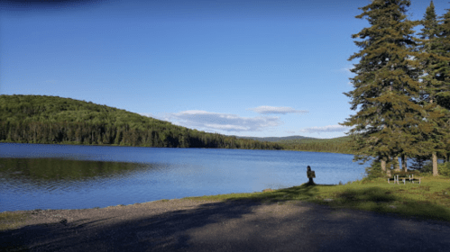 A serene lake surrounded by green hills and trees, with a person standing by the water's edge under a clear blue sky.