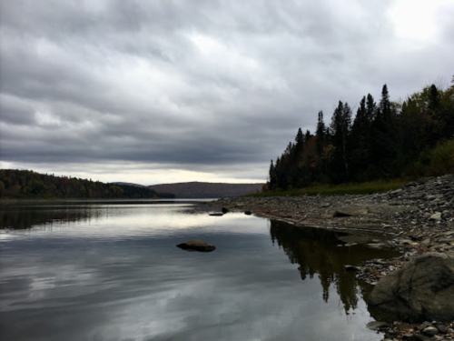 A calm river reflects cloudy skies, bordered by rocky shores and trees in the distance.