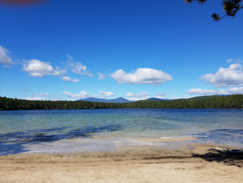 A serene lake with clear blue water, sandy shore, and distant mountains under a bright blue sky with fluffy clouds.