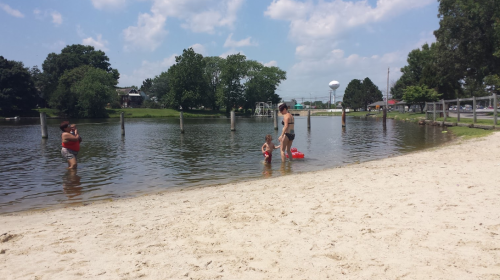 A sandy beach by a calm lake, with people wading in the water and trees in the background under a blue sky.