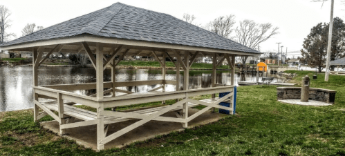 A wooden gazebo by a calm pond, with grassy areas and a stone feature nearby. Trees line the background.