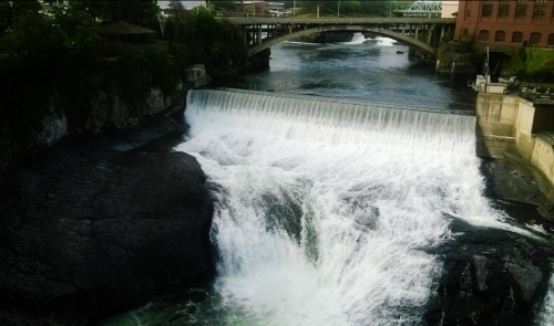 A waterfall cascades over rocks into a river, with a bridge and buildings in the background.