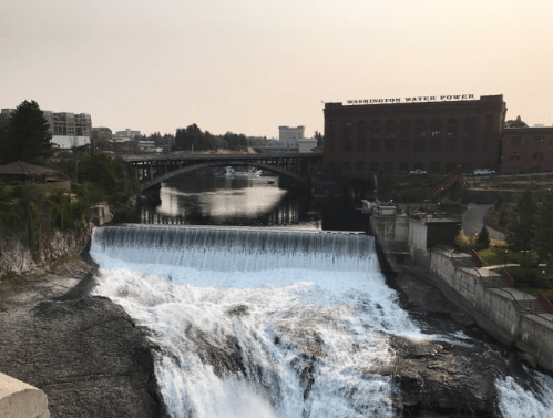 A waterfall cascades over a dam, with a historic building and city skyline in the background under a hazy sky.