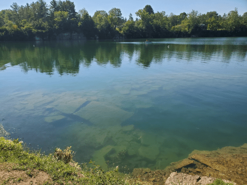 A serene lake with clear water reflecting trees, showing submerged rocks and a person kayaking in the distance.