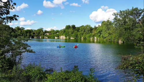 Two kayakers paddle on a calm lake surrounded by lush greenery and blue skies with fluffy clouds.