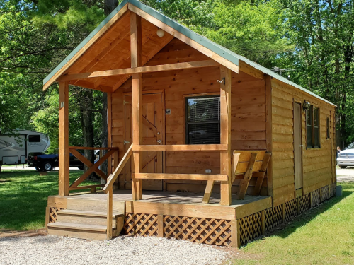 A small wooden cabin with a porch, surrounded by greenery and parked vehicles in a campsite setting.