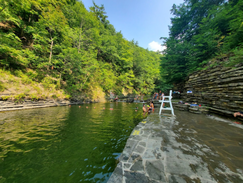 A serene swimming area surrounded by lush greenery and rocky cliffs, with people enjoying the water on a sunny day.