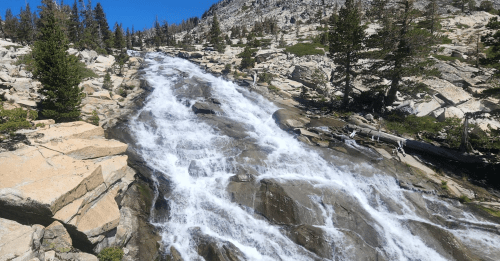 A cascading waterfall flows over rocky terrain, surrounded by trees and a clear blue sky.
