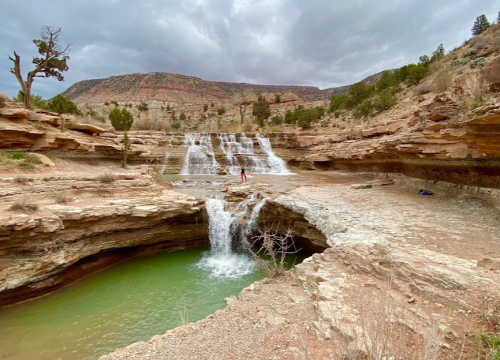 A scenic view of a waterfall cascading over rocky terrain into a green pool, surrounded by dry landscape and hills.