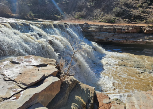 A waterfall cascades over rocky ledges, surrounded by dry terrain and sparse vegetation under a clear blue sky.