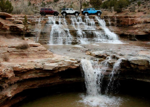 Three off-road vehicles parked near a waterfall cascading over rocky terrain in a desert landscape.