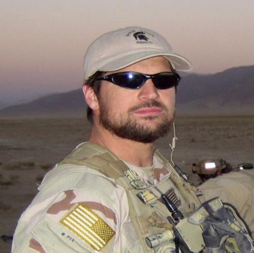 A man in military gear and sunglasses stands in a desert landscape, wearing a cap and a tactical vest.