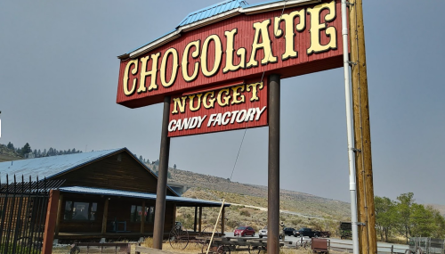 Sign for "Chocolate Nugget Candy Factory" with a rustic building and mountainous background.