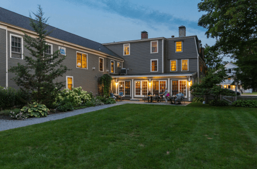 A cozy backyard with a lawn, patio furniture, and people enjoying the evening near a charming house at dusk.