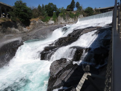 A cascading waterfall flows over rocky terrain, surrounded by greenery and a bridge in the background.