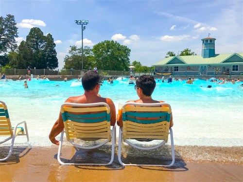 Two people sit in lounge chairs by a wave pool, enjoying the sunny day while others play in the water.