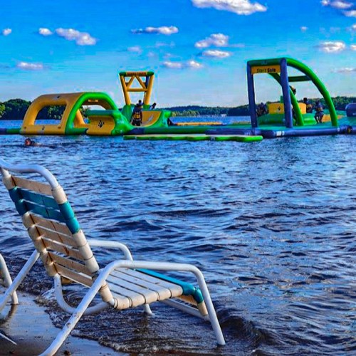 A beach chair sits on the shore, overlooking a colorful inflatable water park in a lake under a blue sky.