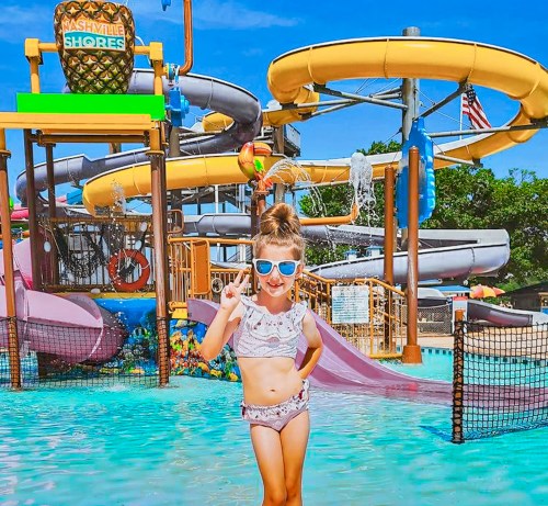 A young girl in sunglasses poses playfully in a water park with colorful slides and a splash pool in the background.