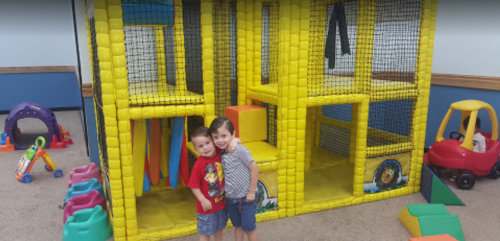 Two children smile in front of a colorful indoor play structure made of soft blocks, with various toys around them.
