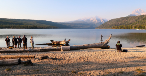A serene lakeside scene with photographers capturing the landscape, surrounded by logs and mountains in the background.