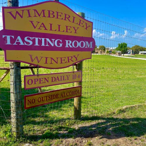 Sign for Wimberley Valley Tasting Room Winery, stating open hours and no outside alcohol allowed, with a clear blue sky.