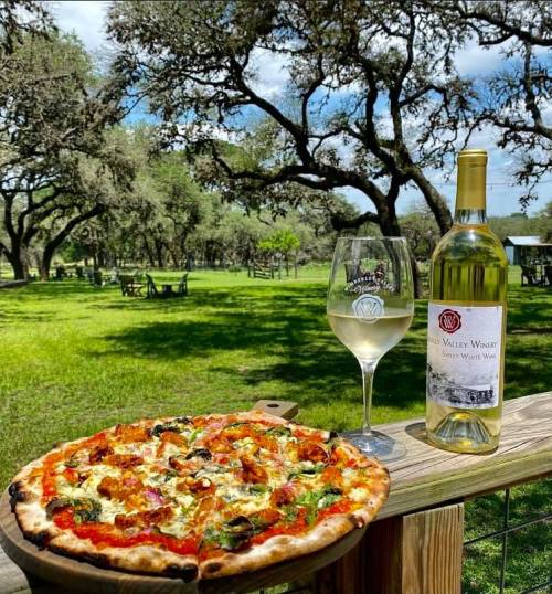 A bottle of white wine and a glass sit beside a pizza on a wooden table, with trees and a sunny outdoor setting in the background.