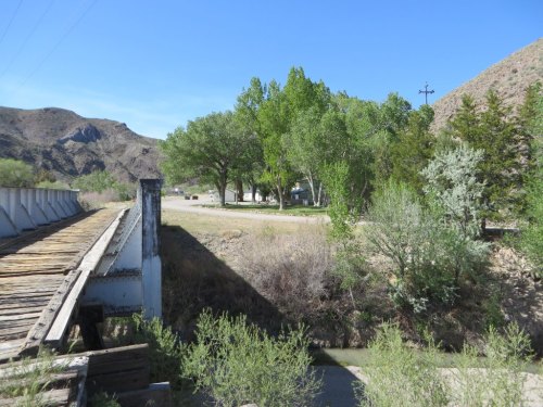 A wooden bridge leads to a grassy area with trees and mountains in the background under a clear blue sky.