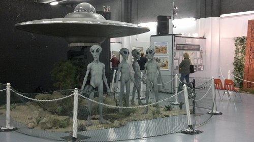 A display featuring a UFO and four alien figures in a museum setting, surrounded by a rope barrier.