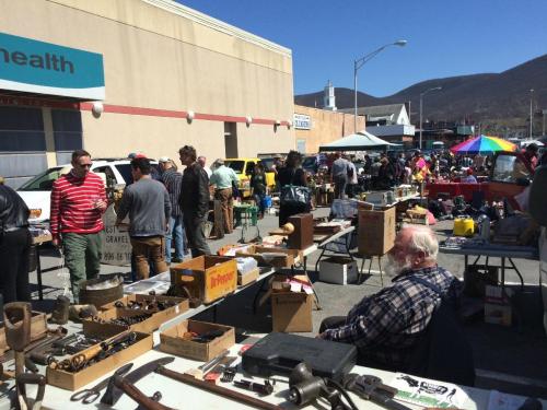 A bustling outdoor flea market with vendors and shoppers browsing various items and tools on tables.