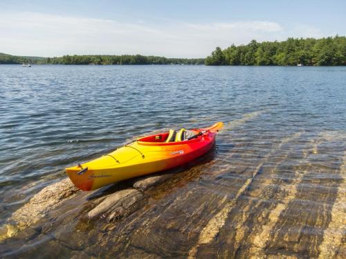 A bright yellow and red kayak rests on a rocky shore by a calm lake, surrounded by green trees and blue sky.