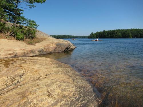 A serene lakeside scene with rocky shores, clear water, and a person kayaking in the distance under a blue sky.
