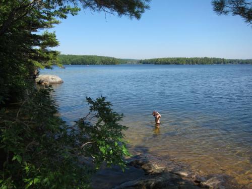A person wades in a clear lake surrounded by trees and rocky shores under a bright blue sky.