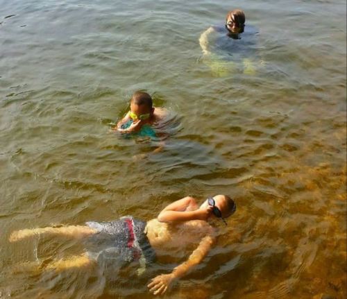 Three children play in shallow water; one floats on their back while the others swim nearby, all wearing goggles.