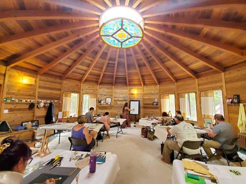 A group of people in a circular wooden room, engaged in a workshop with a presenter at the front and art supplies on tables.