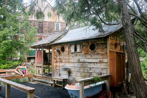 A rustic wooden cabin surrounded by trees, with a person relaxing in a bathtub and another on a nearby bench.