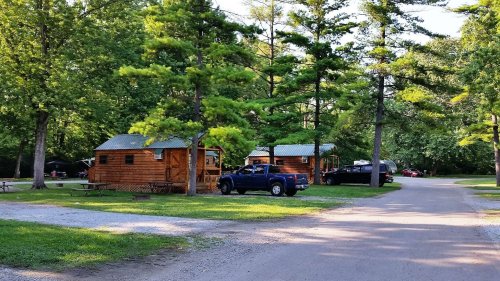 Two wooden cabins surrounded by trees in a campground, with a truck parked nearby on a gravel road.