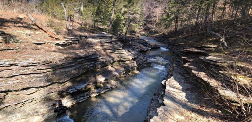 A serene river flows through layered rock formations, surrounded by trees in a peaceful natural setting.