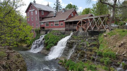 A red historic mill beside a flowing river with two small waterfalls and lush greenery surrounding the area.
