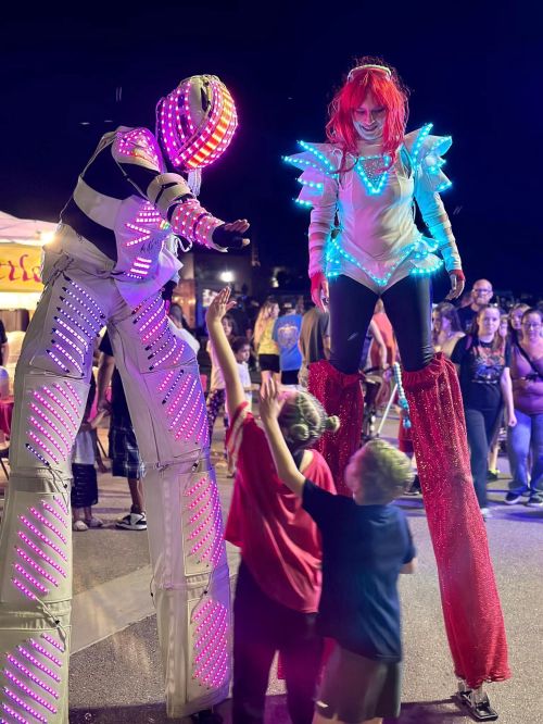 Two performers on stilts with colorful LED lights interact with children at a lively night festival.