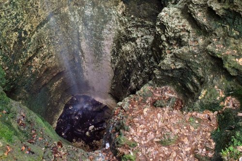 A deep, moss-covered pit surrounded by rocky terrain and scattered leaves, with a faint mist rising from the bottom.