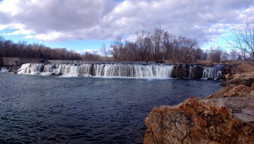 A scenic view of a waterfall cascading over rocks, surrounded by trees and a cloudy sky.