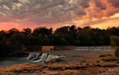 A serene waterfall cascades over rocks under a colorful sunset sky, surrounded by lush trees and clouds.