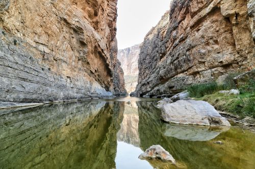 A narrow canyon with steep rock walls reflecting in calm water below. Lush greenery peeks from the rocky edges.